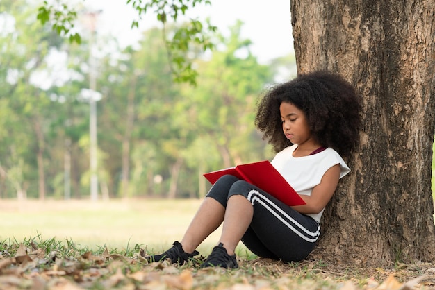 Ragazza afroamericana con capelli ricci che legge un libro sotto l'albero nel parco. concetto di educazione all'aperto.