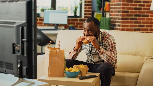 African american guy having fun eating burgers and drinking beer, watching comedy movie on television. Male adult enjoying fast food delivery and alcohol, watch tv show in living room.