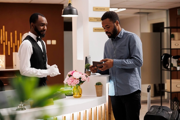 African American guy hotel guest showing his passport to receptionist standing behind reception counter. Traveler with suitcase checking in at front desk upon arrival. People and traveling