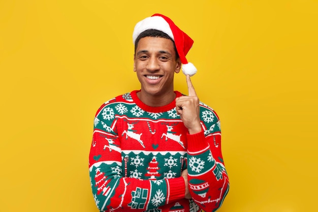 African american guy in New Year's clothes and santa hat smiles on yellow isolated background