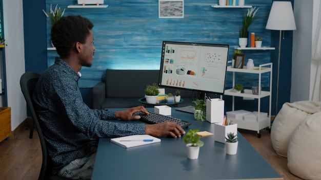 African american guy working from home, taking notes on notepad while remote working on computer from living room