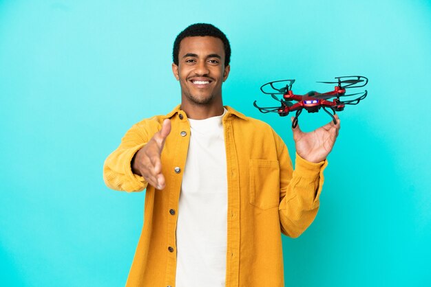 African american handsome man holding a drone over isolated blue background shaking hands for closing a good deal