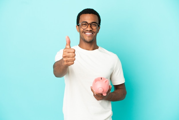 African American handsome man holding a piggybank over isolated blue background with thumbs up because something good has happened