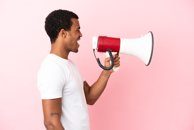 Афро-американский красавец на изолированном розовом фоне кричит в мегафон, чтобы объявить что-то в боковом положении