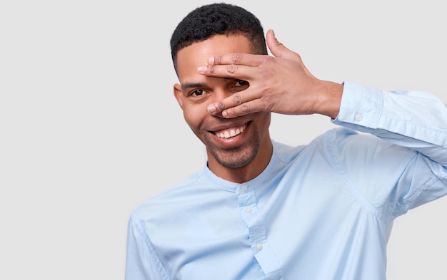 African American handsome young man hiding his face with palm and show his eye Studio portrait of young male covering his face by hand wearing blue shirt and looking at camera posing on white wall