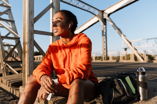 african american happy woman in sportswear using earpods and holding cellphone while sitting on old bridge