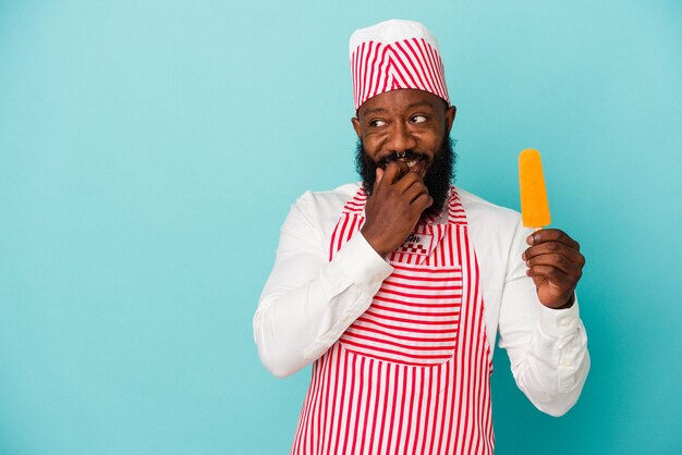 African american ice cream maker man holding an ice cream isolated on blue wall relaxed thinking about something looking at a copy space.