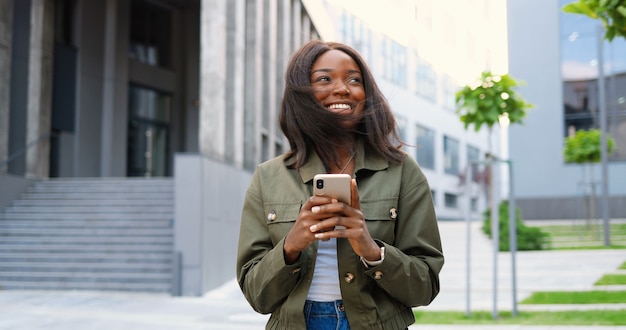 スマートフォンをタップまたはスクロールし、街の通りに立っているアフリカ系アメリカ人の楽しい若いスタイリッシュな女性。携帯電話と笑顔で美しい幸せな女性のテキストメッセージ。外側。