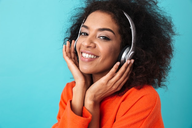 African american lachende vrouw in oranje shirt luisteren naar muziek via draadloze koptelefoon, geïsoleerd over blauwe muur