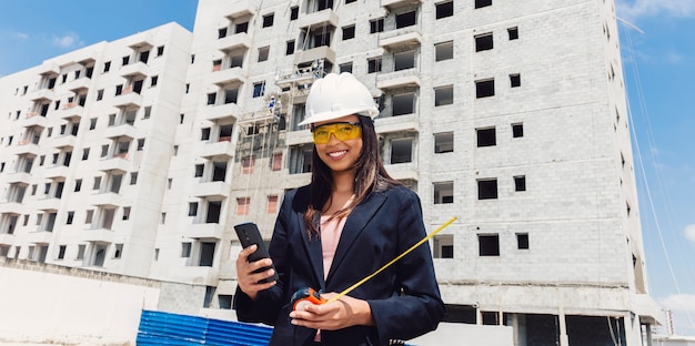 Photo african american lady in safety helmet with smartphone and measuring tape near building under construction