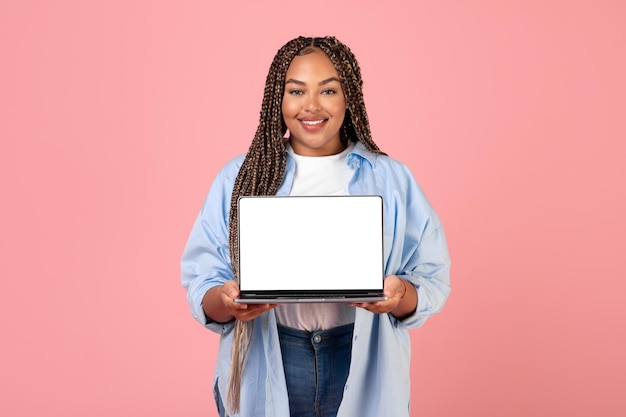 Афроамериканка показывает пустой экран ноутбука на розовом фоне