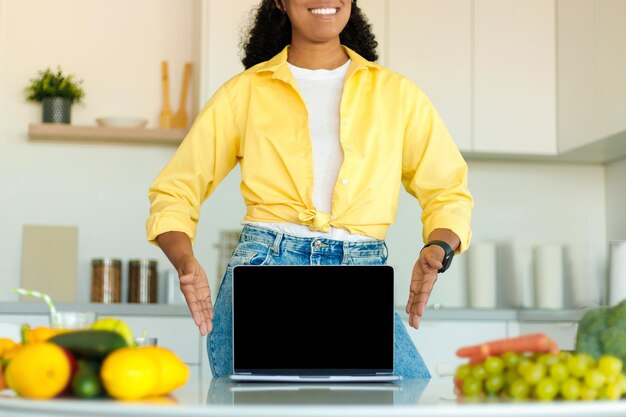 African american lady showing laptop with blank screen at table\
with organic vegetables at kitchen interior mockup