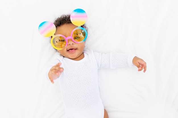 An African-American little child in funny glasses lies on a white bed and smiles