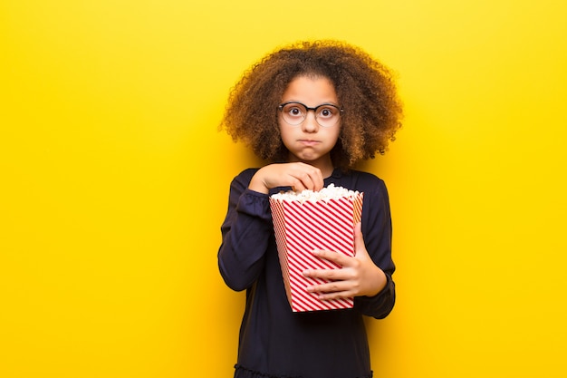 Афро-американская маленькая девочка против плоской стены, держащей ведро попкорна
