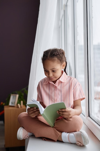 African american little girl sitting on window and reading book