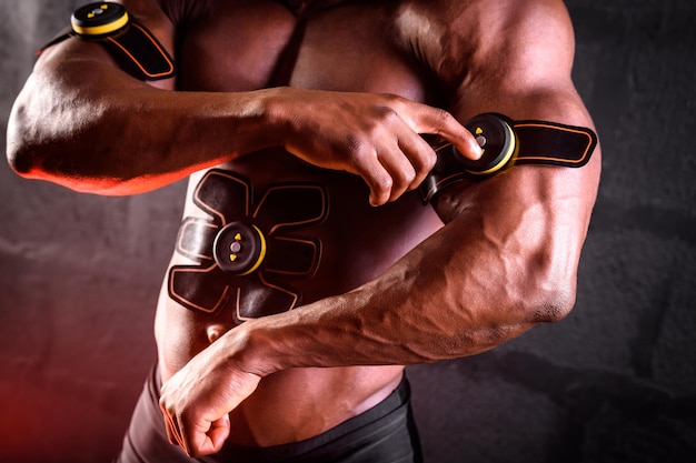 African american male bodybuilder uses electronic myostimulator to train biceps hands and abdominal muscles on a black background