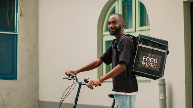 アフリカ系アメリカ人の男性の宅配便業者が自転車に乗って顧客に食事のパッケージを渡し、フロントドアにバックパックを運んでいます。自転車を操作し、レストランのテイクアウト食品を配達する配達員。