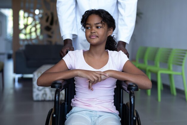 African american male doctor pushing smiling mixed race girl in wheelchair through hospital corridor