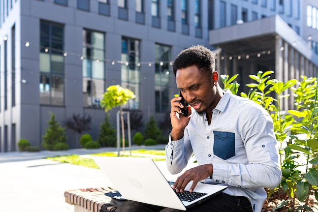 African american male freelancer working remotely online broker\
with exchanges stocks talking on mobile phone and laptop merchant\
seller. male in city park a bench modern urban background business\
man