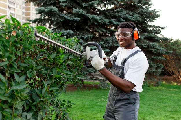 African american male garden worker in uniform trims bushes with electric tool