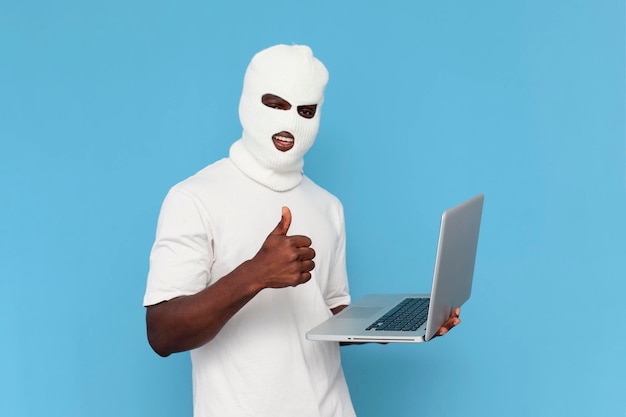 흰색 발라클라바를 입은 아프리카계 미국인 남성 해커는 마스크를 쓴 파란색 배경의 노트북을 사용합니다.