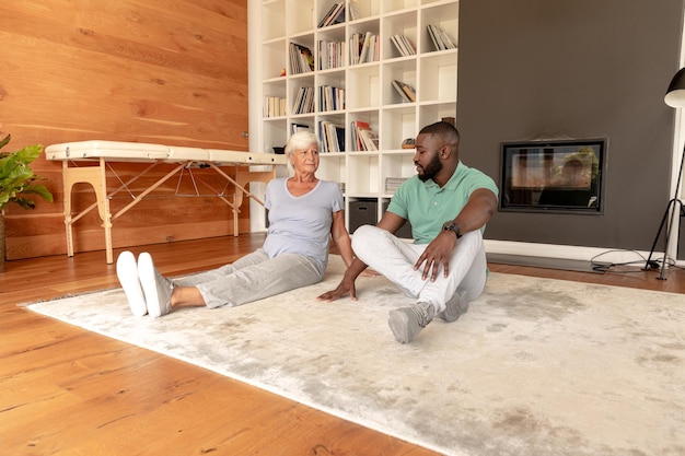 Африканский американский физиотерапевт и кавказская пожилая женщина сидят дома на полу