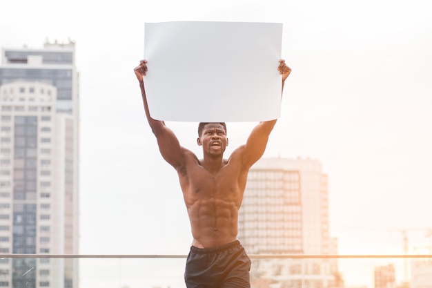 African American male protester holds a blank poster and shouts against the backdrop of the city