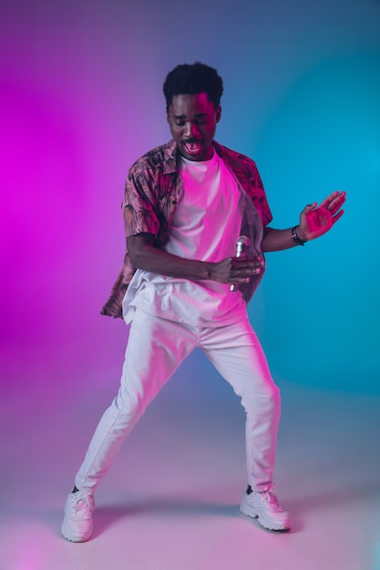 Ritratto di cantante maschio afroamericano isolato su sfondo sfumato studio in luce al neon. bellissimo modello maschile con microfono. concetto di emozioni umane, espressione facciale, pubblicità, musica, arte.