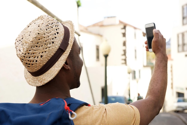Photo behind of african american male tourist with bag and hat taking selfie