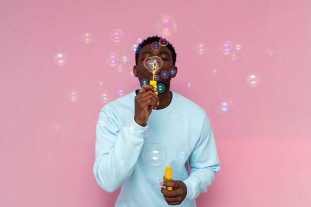 african american man in blue sweater inflates soap bubbles on pink isolated background