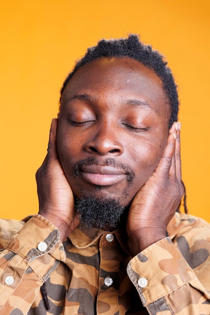 African american man covering ears with palms, doing three wise monkeys gesture in front of camera over yellow background. Person with serious expression not listening to noise and not speaking