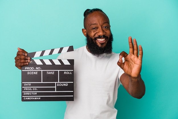 African american man holding clapperboard isolated on blue background cheerful and confident showing ok gesture.