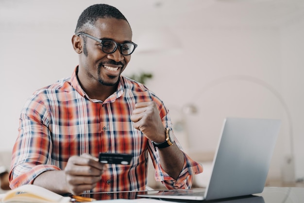 African american man holding credit card uses online banking service at laptop makes yes gesture