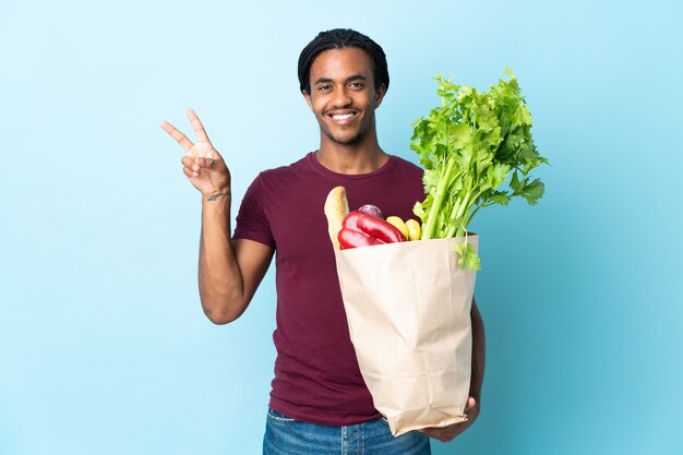 青い笑顔と勝利のサインを示す食料品の買い物袋を保持しているアフリカ系アメリカ人の男