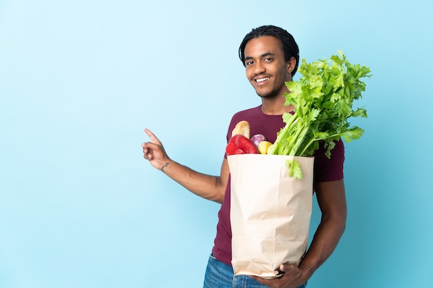 African american man holding a grocery shopping bag isolated on
blue background pointing back