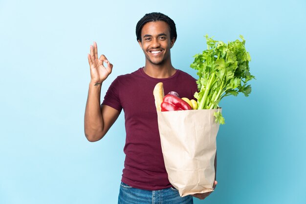 Photo african american man holding a grocery shopping bag isolated on blue background showing ok sign with fingers