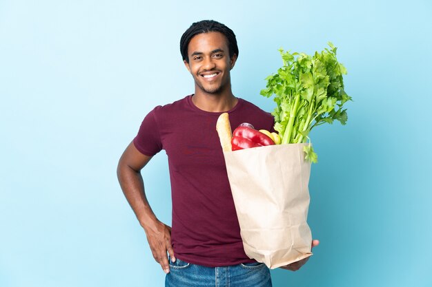 African American man holding a grocery shopping bag isolated on blue posing with arms at hip and smiling