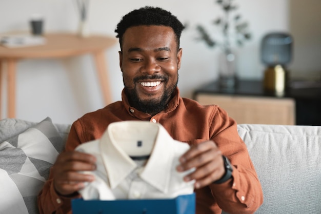 African american man holding new shirt unpacking shopper bag indoors