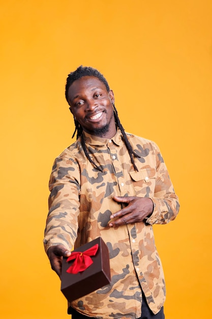 African american man holding wrapped gift box for girlfriend, posing for valentine s day in studio over yellow background. Stylish smiling person feeling confident during love holiday.