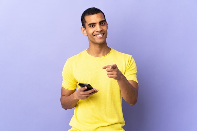 African American man over isolated purple background using mobile phone points finger at you