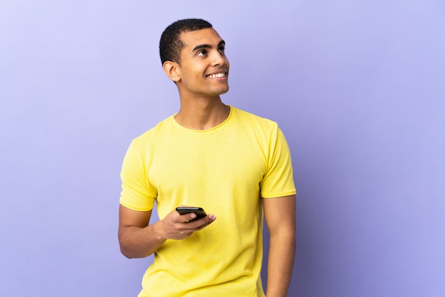 African American man over isolated purple wall using mobile phone laughing and looking up