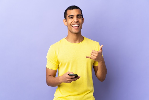 African American man over isolated purple wall using mobile phone making phone gesture