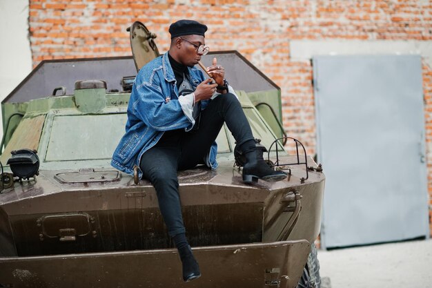 African american man in jeans jacket beret and eyeglasses lights a cigar and posed against btr military armored vehicle