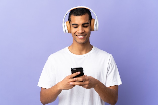 African American man over geïsoleerde paarse muur luisteren muziek en op zoek naar mobiel