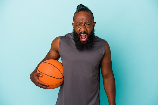 Афро-американский мужчина играет в баскетбол, изолированные на синей стене, кричит очень сердито и агрессивно.