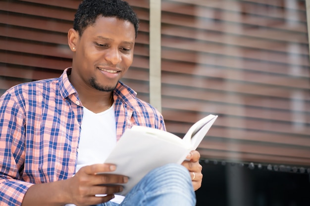 아프리카 계 미국인 남자가 편안하고 거리에 상점 창에 앉아있는 동안 책을 읽고.