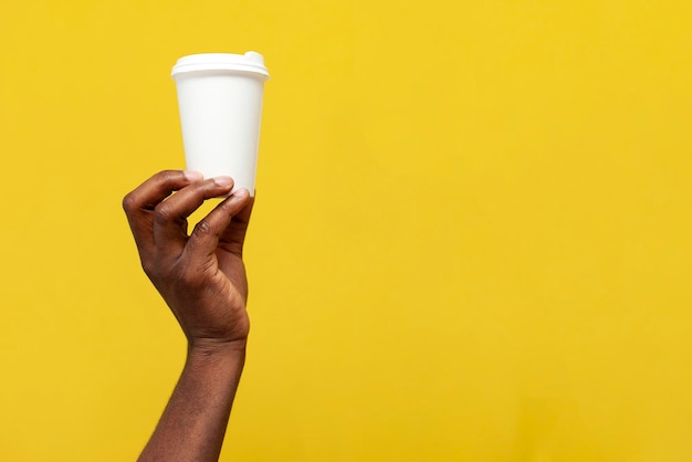 African american man's hand holds white paper cup of coffee on yellow isolated background