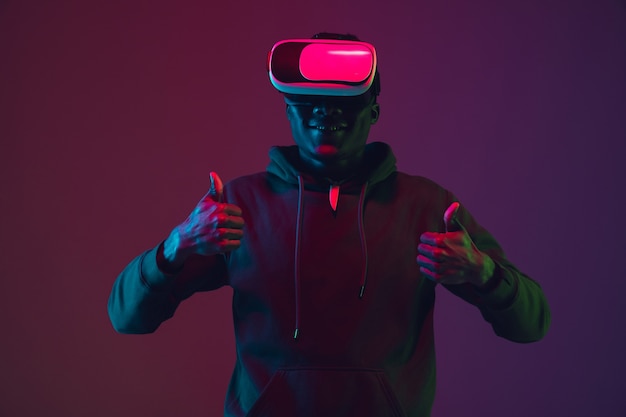 African-american man's portrait in VR-headset isolated on gradient studio background in neon light. Beautiful male model. Concept of human emotions, facial expression, sales, ad, inclusion, tech.