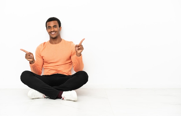 African American man sitting on the floor over isolated copyspace background pointing finger to the laterals and happy