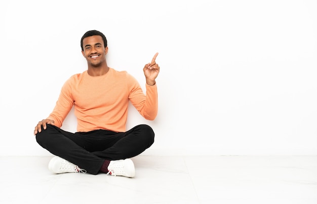 African American man sitting on the floor over isolated copyspace background showing and lifting a finger in sign of the best